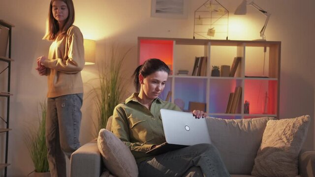 Family Secrets. Conspirator Woman. Digital World. Curious Daughter Looking At Laptop Of Suspicious Mother Passing By Her Sitting Sofa In Light Home Interior.