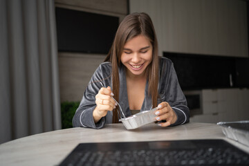 Happy woman enjoying a delicious takeaway lunch at home. A beautiful girl sits at a table and eats from a food container. Food delivery service concept. makes a video call on a laptop.