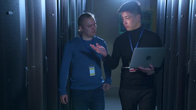 Two data engineers talking and using laptop when doing diagnostics in server room spbas. Young men talk and inspect hardware shelves, hold device in hand and walk in dark database corridor