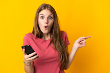 Young woman using mobile phone isolated on yellow background surprised and pointing side