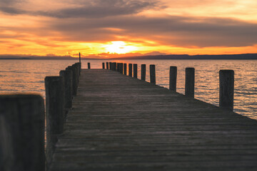 Wooden footbridge with pilings at sunset on Lake Neusiedl. The orange sun reflects in the waves of the water. The cloudy sky makes the photo very atmospheric.