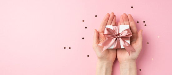 First person top view photo of valentine's day decorations woman's hands holding small white giftbox with pink ribbon bow on palms and confetti on isolated pastel pink background with empty space