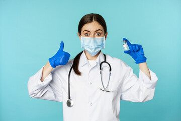 Portrait of smiling doctor, medical personel in face medical mask and rubber gloves, showing thumbs up and coronavirus, omicron vaccine, standing over blue background - obrazy, fototapety, plakaty