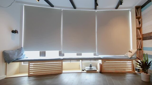 Video of motorized roller shades in the interior. Automatic roller blinds beige color open up on big glass windows. Remote Control Shades are above the windowsill with pillows. Summer. 