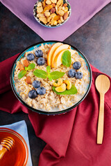 Oatmeal porridge with yogurt, berries and nuts on burgundy napkin. Bowl with oatmeal, blueberries, apples and honey on a dark background. Healthy breakfast. Top view