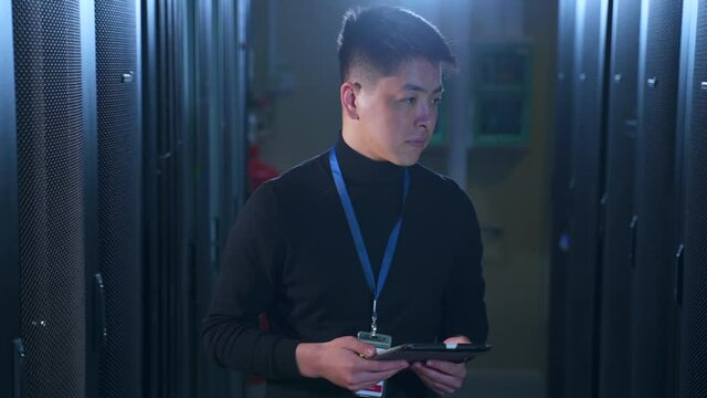 Young male engineer using device and checking equipment, walking in data office spbas. Close-up view of Asian man inspects hardware racks and holds tablet in hand, does analytical work and walks in