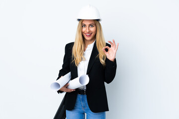 Young architect woman with helmet and holding blueprints over isolated white background showing an ok sign with fingers