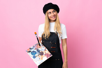 Young artist woman holding a palette over isolated pink background laughing and looking up