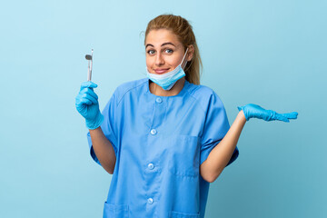 Young woman dentist holding tools over isolated blue background having doubts with confuse face...