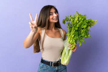 Young woman holding a celery isolated on purple background smiling and showing victory sign