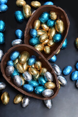 Pile of colourful foil wrapped chocolate easter eggs in blue, silver, and gold with two halves of a large brown milk chocolate egg in the middle and mini eggs inside.