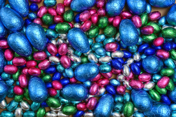Fototapeta na wymiar Large & small pink, blue, green and silver foil wrapped chocolate easter eggs, against a pale wood background.