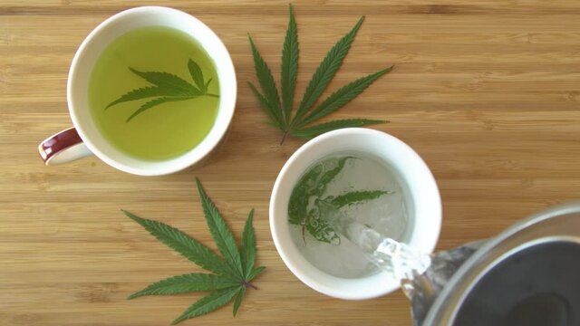 SLOW MOTION, TOP DOWN, CLOSE UP: Hot water gets poured over a homegrown marijuana leaf inside of a white teacup sitting on the wooden dining table. Person is preparing therapeutical medicinal weed tea