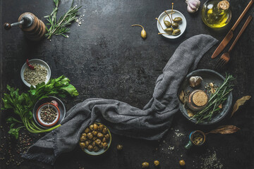 Mediterranean food background with olives, oil, parsley, rosemary, caper and kitchen utensils on black concrete kitchen table. Greek cuisine with tasty ingredients. Top view with copy space. Frame