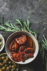 Sun-dried tomatoes in olive oil in white bowl on dark rustic kitchen table with rosemary and...