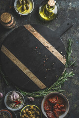 Various Mediterranean ingredients around empty dark circle cutting board : sun-dried tomatoes, herbs, capers, olives, olive oil and garlic. Preparing homemade antipasti plate. Top view. Frame