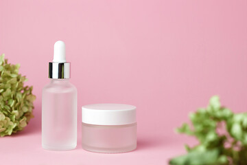 Two white glass cosmetic jars for cream and serum on a pink background with green foliage. Cosmetology and beauty. Self care. Model for cosmetics.