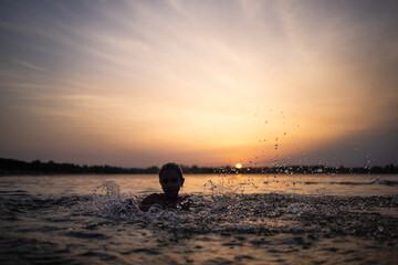 A girl plays with water in the lake and splashes it to the sides against the backdrop of a sunset