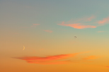 Sunset with silhouette of paragliders flying over the orange sky with bright crescent moon