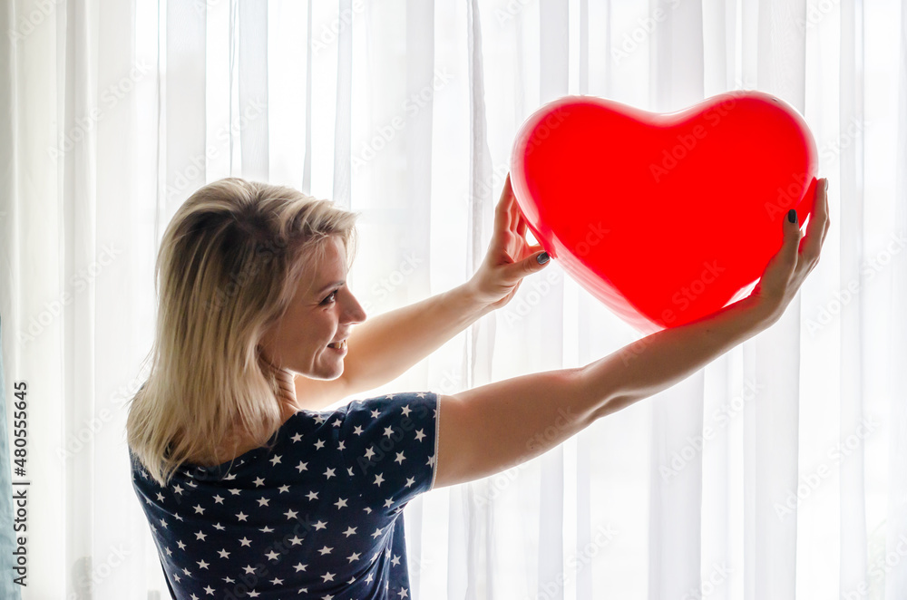 Wall mural young woman holding red heart shaped baloon against the window at home. celebrating valentines day, 