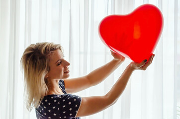 Young woman holding red heart shaped balloon against the window at home. Celebrating valentines day, wedding and love day, women's day, World Heart Day, birthday. Silhouetted against the sun indoor.