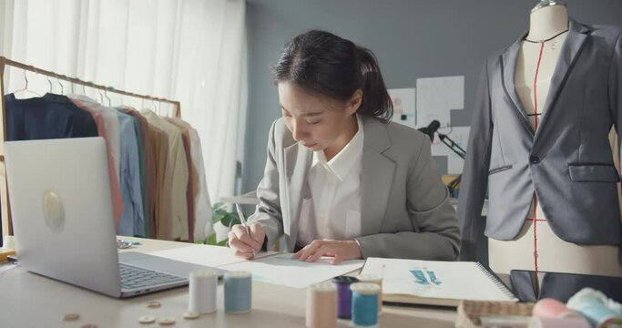 Professional cheerful Asia businesswoman designer sitting on table with laptop computer draw design dress fashion new collection on sketchbook work project at studio workplace. Successful business.