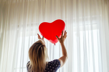Young woman holding red heart shaped balloon against the window at home. Celebrating valentines...