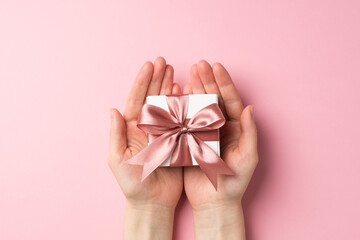 First person top view photo of valentine's day decorations female hands holding small white giftbox with pink ribbon bow on palms on isolated pastel pink background