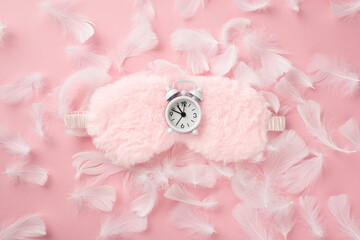 Top view photo of white alarm clock on pink fluffy sleeping mask and feathers on isolated pastel pink background