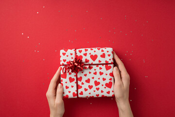 First person top view photo of saint valentine's day decor sequins female hands taking giftbox in white wrapping paper with heart pattern and red star bow on isolated red background with blank space