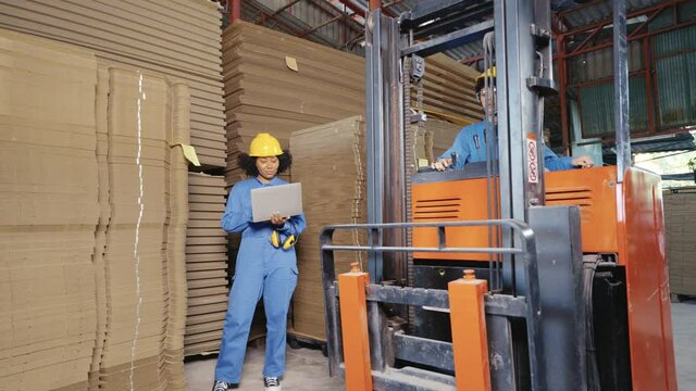 Asian female warehouse worker in blue uniform, helmet, and goggles driving forklift truck through female colleague standing using laptop in front of a large stack of flat cardboard boxes in warehouse.