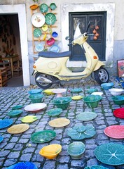 Vertical of yellow scooter parked in front of pottery workshop with colorful plates and bowls  displayed on cobblestone street  at Feidera da Ladra (Campo de Santa Clara) market in Lisbon, Portugal.