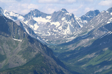 Obraz premium Rocky peaks of a high-mountain ridge on the Kara-Turek pass in Altai with snow, a glacier and a small lake in summer, rocky mountain slopes on the right and left, a sky with clouds, sunny