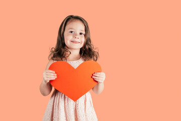 Portrait of little girl with long curly hair hold in hands red paper heart smiling isolated on peach background. Family holidays, Valentines day, birthday, Mother's day concept. Copy space.