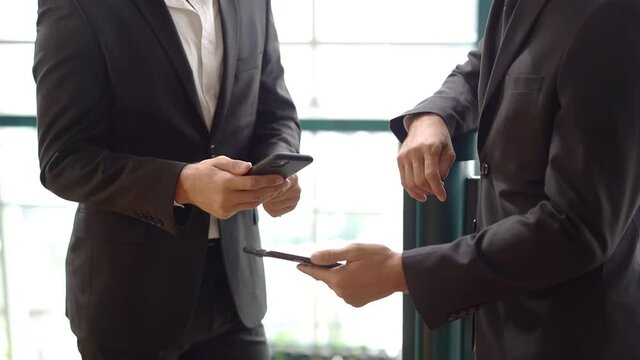 Cropped view of two smart businessmen in a black suits standing on the office corridor using mobile phone to make a contact online by scanning qr code from the other for adding friend on social media.