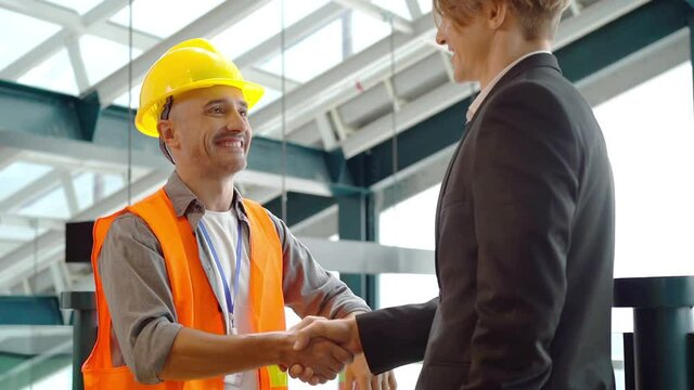A middle-aged Latino male engineer with a thin beard in a reflective vest and helmet, smiling shaking hands and talking to a young handsome Caucasian businessman in a suit at a construction site.
