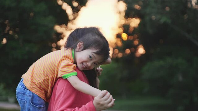 happy asian mother play with baby girl outdoor in the sunset park lovely chinese kid lying on her mother's back mom and daughter relax lifestyles outdoors slow motion 4k clip