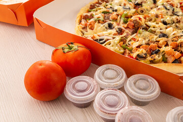cardboard packaging with a delicious pizza on a table next to some tomatoes decorating and containers with oregano, fast food and lifestyle