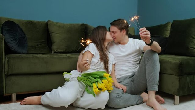 Happy young couple celebrating love with a sparklers in hands, woman have a bouquet of yellow tulips from her husband, kissing, romance, anniversaries.