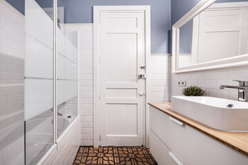White bathroom with shower and glass partition, porcelain sink on wooden countertop and hydraulic tile floors
