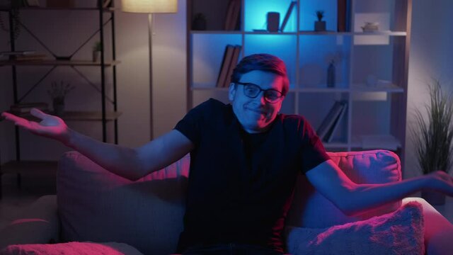 Shrugging hands. Confused man. Meme expression. Clueless funny guy in spectacles raising shaking hands sitting sofa in dark neon light home interior looped.