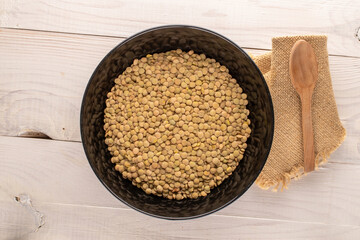 Uncooked organic green lentils in a black ceramic plate and a wooden spoon on a wooden table, macro, top view.