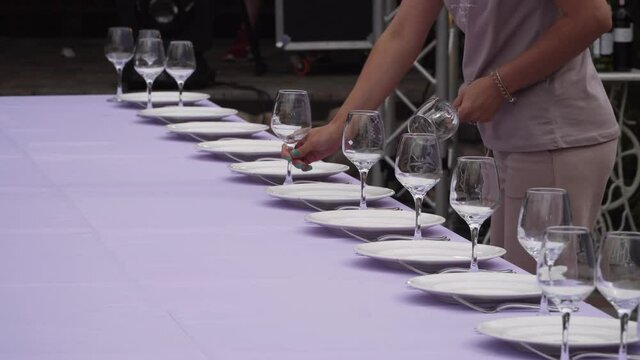 Unrecognizable caucasian woman arranges clean, empty glasses next to plates on banquet festive table with purple tablecloth. Waiter prepares table for party outdoor. Cutlery is next to the plates.