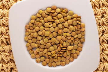 Uncooked organic green lentils in a white plate on a straw mat, macro, top view.