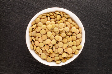 Uncooked organic green lentils in a white saucer on a slate stone, macro, top view.