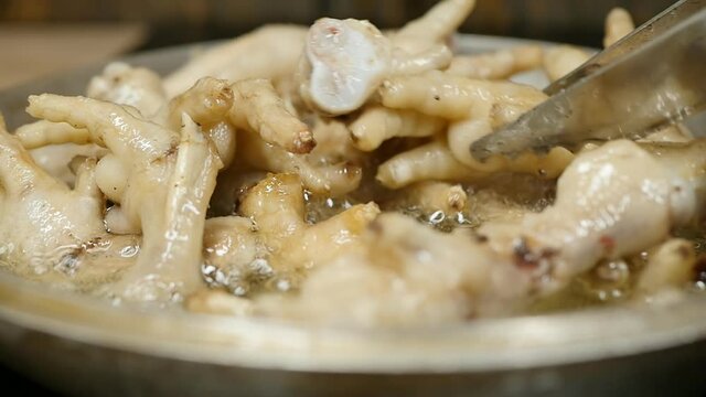 the cook cooks chicken feet in hot vegetable oil in a frying pan flips the legs with steel grill tongs. Slow motion bubbles of boiling fat