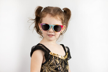 Little cheerful girl with two ponytails and a black and gold dress in sunglasses. Studio photo on a white background of a half-turned sad child