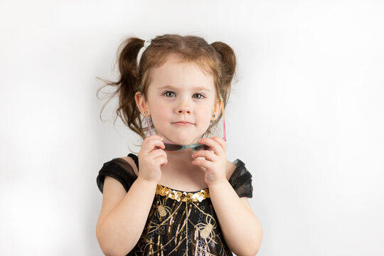 A little serious girl with two ponytails and a black and gold dress takes off her sunglasses. Studio photo of a child on a white background