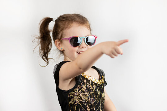 Little cheerful girl with two ponytails and a black and gold dress in sunglasses. Studio photo on a white background of a sad child pointing with his index finger
