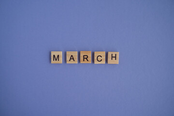The name of the month MARCH is written on wooden isolated cubes. Very Peri Color of the Year 2022 background, copy space. Top view of wooden blocks with letters. Step by step, step 3.
